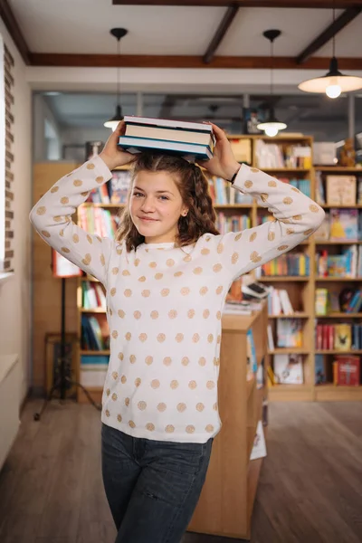 Beautiful girl is holding stack of books while standing among books in the bookshop — Stok fotoğraf
