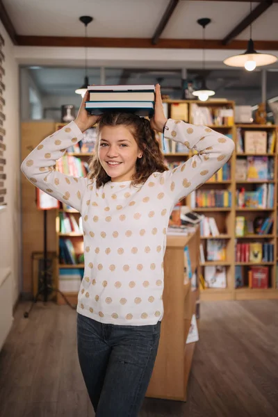 Beautiful girl is holding stack of books while standing among books in the bookshop —  Fotos de Stock
