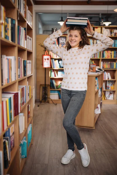 Beautiful girl is holding stack of books while standing among books in the bookshop — стоковое фото