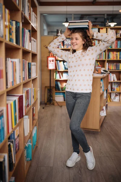 Beautiful girl is holding stack of books while standing among books in the bookshop — Stockfoto