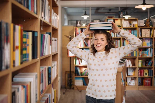 Beautiful girl is holding stack of books while standing among books in the bookshop — Stock Fotó