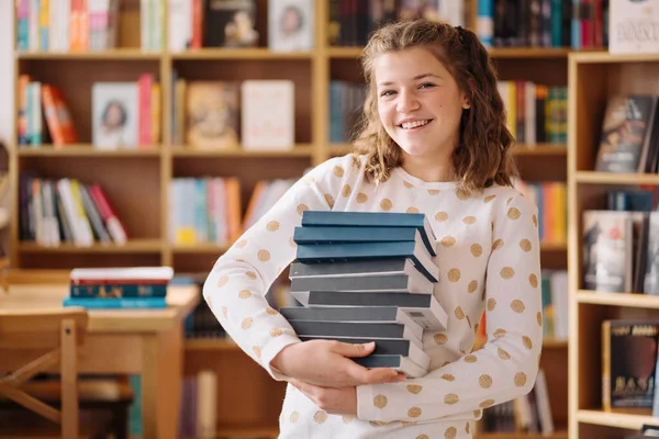 Beautiful girl is holding stack of books while standing among books in the bookshop —  Fotos de Stock