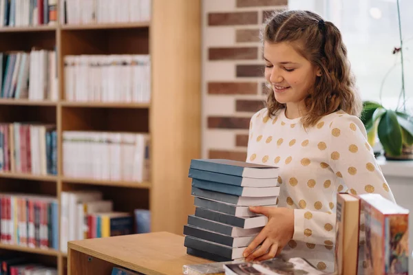 Beautiful girl is holding stack of books while standing among books in the bookshop — Stockfoto