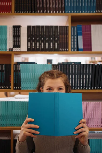 背景に本棚のある本の後ろに顔を隠す少女 — ストック写真