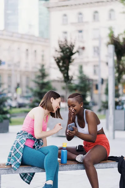 Two multiethnic girls relaxing after exercise session, outdoor communicating and using smartphone — Stock Photo, Image