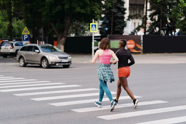 Dwie wielonarodowe dziewczyny biegające po ulicach miasta, porozumiewające się na świeżym powietrzu. — Zdjęcie stockowe