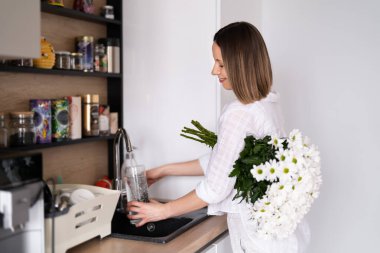 Çiçekleri yerleştirmek için mutfak lavabosundaki vazoya su döken güzel kadın.