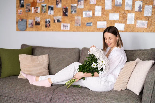Güzel bir kadın evdeki kanepede otururken beyaz çiçeklerden hoşlanır. — Stok fotoğraf