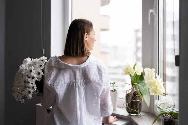 Νεαρή γυναίκα κοιτάζει έξω από το παράθυρο την άνοιξη κρατώντας λευκά λουλούδια — Φωτογραφία Αρχείου