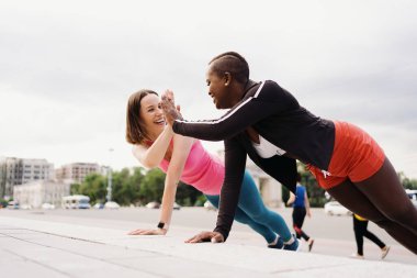 Spor kıyafetleriyle gülümseyen iki genç kadın şehir meydanında Plank egzersizi yapıyor..
