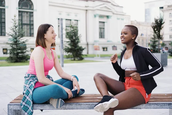 在城市的长椅上 穿着运动服的快乐的微笑的朋友们在公园里闲聊着 多族裔妇女有一个健身休息时间 — 图库照片