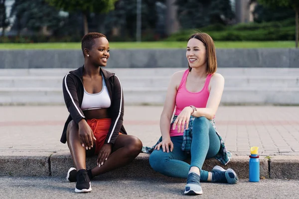 穿着运动服的快乐微笑的朋友们坐在城市里口齿不清 多族裔妇女有一个健身休息时间 — 图库照片
