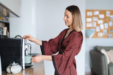 Evde kahve yapmak için kahve makinesi kullanan bir kadın. Evde latte hazırlarken ipek bornoz giyen bir kadın.