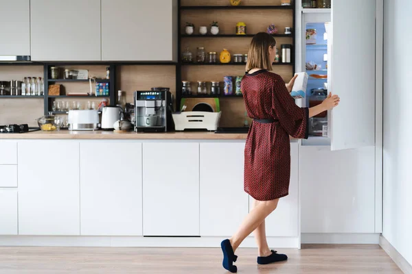 自宅で冷蔵庫で食べ物を探している空腹の女性が そこにはあまりありません 白いキッチン家具 ホームウェア 赤いシルクのローブ — ストック写真
