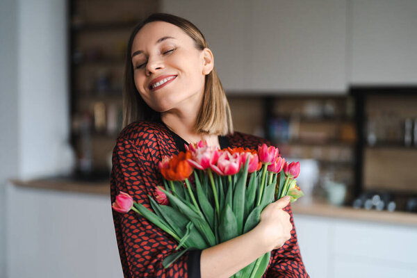 Happy woman enjoy bouquet of tulips. Housewife enjoying a bunch of flowers and interior of kitchen. Sweet home. Allergy free
