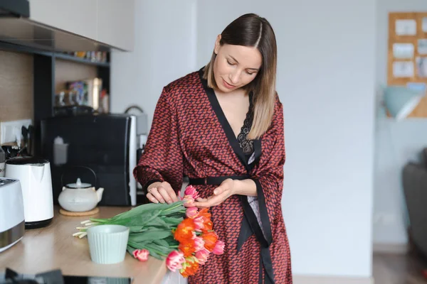 快乐的女人在厨房喝咖啡 身穿丝绸长袍 欣赏鲜花 — 图库照片