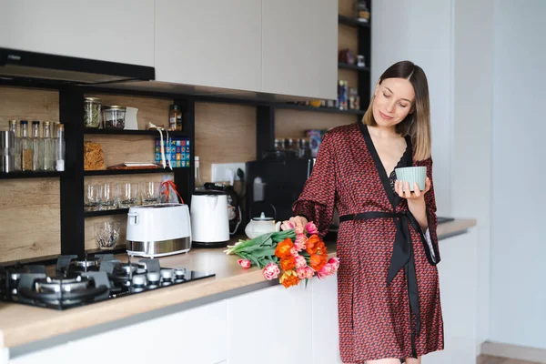 Happy Woman having a coffee at home in the kitchen wearing a silk robe while enjoying fresh flowers