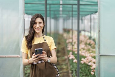 Smiling florist businesswoman using smartphone, wearing yellow t-shirt and brown apron holding phone in hands at the green house