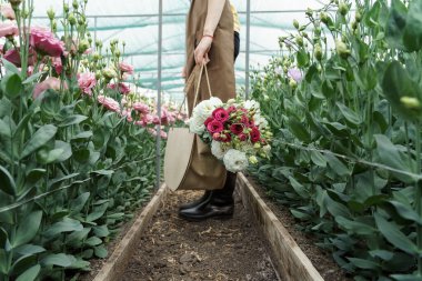 Woman florist walking among flowers in a green house carrying a basket with a fresh bouquet
