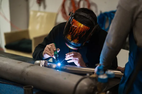 Worker in helmet with flames welding the steel part of a motorbike using an argon welding machine and gloves in a garage