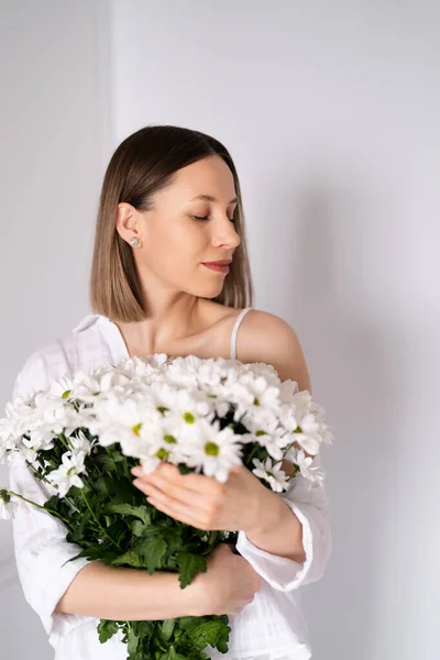 白い壁の背景に白い新鮮な花の花束を保持して若い美しいかわいい甘い素敵な笑顔の女性 — ストック写真