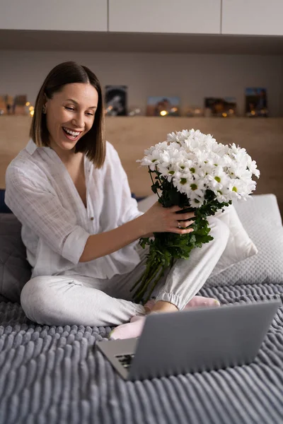 白い花を楽しむ喜びとパジャマを着てベッドの上に座っている若い女性は ラップトップを使用してチャット — ストック写真