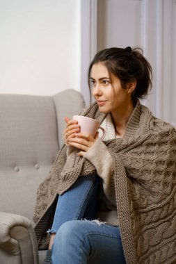 Zeki modaya uygun giyinmiş gülümseyen bayan elinde bir fincan çayla koltukta oturuyor. Gülümsemek, evde rahat bir atmosferde oturmak, güzel tasarım..