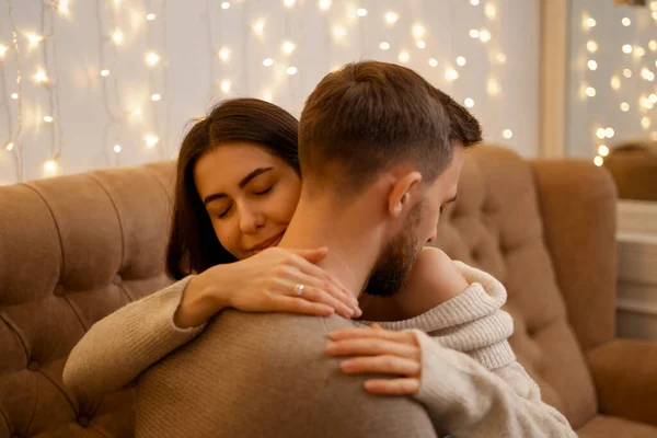 Happy married young couple hugging, sitting on cozy couch together, woman and man enjoying leisure time, relaxing on sofa in living room at home with lights in background