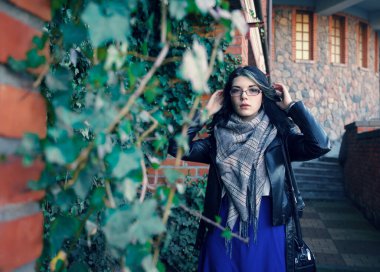 Siyah ceketli ve mavi elbiseli genç esmer kız sonbahar günü eski taş kale duvarının yanında duruyor.