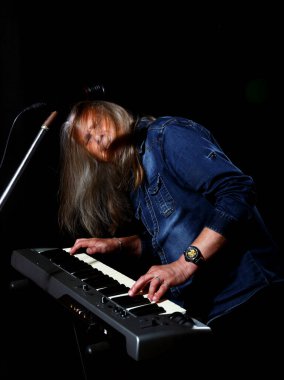 Uzun gri saçlı, mavi kot gömlekli yaşlı bir adam sahnenin anahtarını siyah arka planda çalıyor.