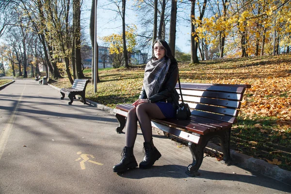 Siyah ceketli ve mavi elbiseli genç esmer kız sonbahar günü şehir parkında bankta oturuyor.