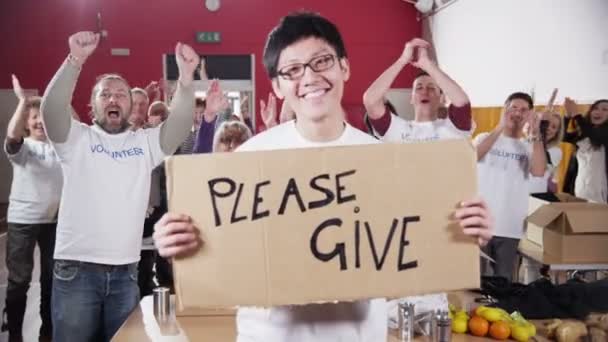 Благодійний працівник тримає знак Будь ласка, дайте знак, як його колеги аплодують — стокове відео