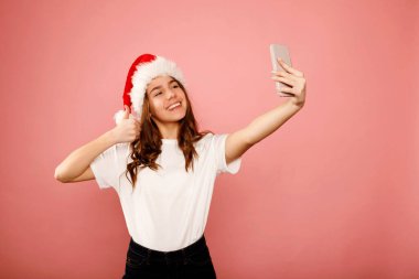 Beyaz tişörtlü ve Noel Baba kırmızı şapkalı mutlu beyaz kız elinde akıllı telefonla pembe arka planda el hareketleriyle selfie çekiyor. Stüdyo portresi. Noel ve Yeni Yıl Tatil konsepti teknolojisi.