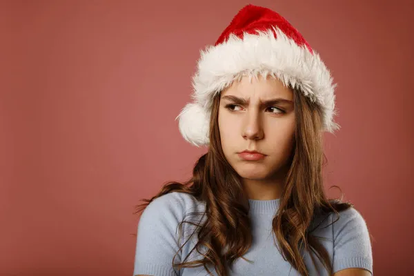 怒气冲冲的高加索女孩少年站在圣塔克洛斯红帽旁边 望着粉红的背景 工作室肖像 反圣诞及新年假期概念并附有复本 — 图库照片