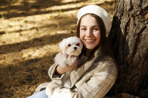 在秋天的公园里 年轻快乐的白人少女头戴法国白帽 身穿保暖外套 牵着并拥抱着她可爱的小白狗的画像 — 图库照片