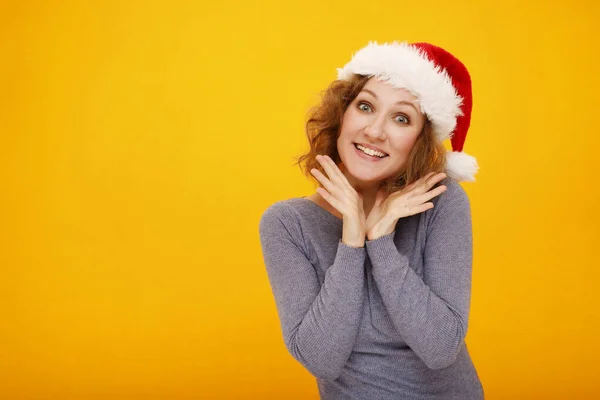 年轻快乐的女人 卷曲的头发 戴着圣诞帽 站在黄色的背景上 笑容满面 看着镜头 手拉手靠近脸 放在你的文字位置上 — 图库照片