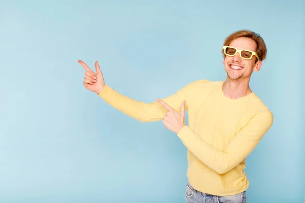 年轻快乐的男人戴着黄色的太阳镜 站在蓝色的背景上 笑容满面 手指头指向侧面 放在你的文字位置上 — 图库照片