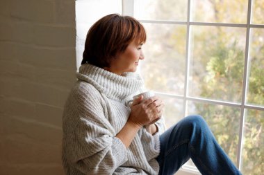 Orta yaşlı, kazaklı, çay ya da kahve içen, pencere kenarında oturan olgun bir kadın. Hygge konsepti.