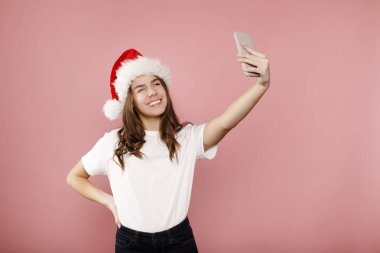 Noel Baba şapkalı kıvırcık saçlı genç kız pembe arka planda duruyor ve selfie çekiyor ve el işaretini gösteriyor. Mutlu yıllar ve Noel konsepti