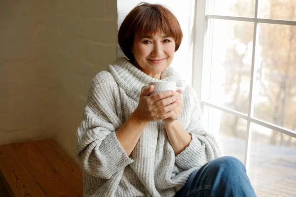 穿着毛衣的中年漂亮的成熟女人坐在窗台上喝茶或喝咖啡 Hygge概念 — 图库照片