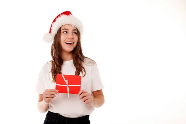 快乐的少女穿着白色T恤 头戴三爪红礼帽 头戴礼品证 站在白色背景上 望着旁边的选择空间 人们的生活方式圣诞节和新年的概念 — 图库照片