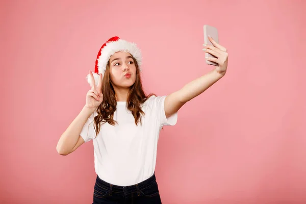 年轻快乐的少女 留着卷曲的头发 戴着圣诞老人的帽子 站在粉红的背景上 两指示意和平 新年快乐 圣诞快乐 放在你的文字位置上 — 图库照片