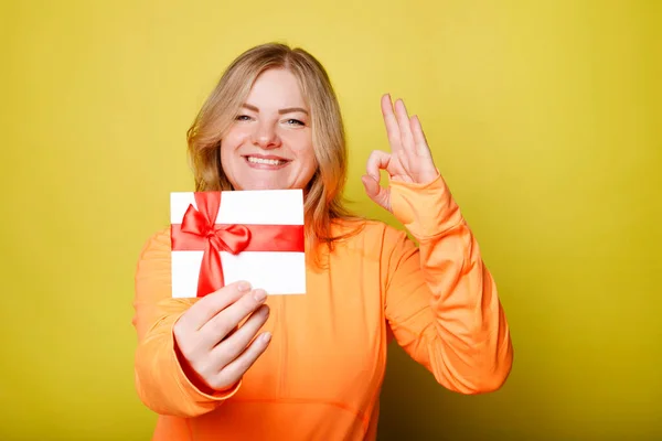 Happy Young Woman Orange Shirt Holds Gift Certificate Isolated Yellow — Fotografia de Stock