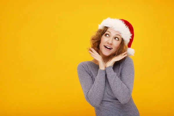 年轻快乐的女人 卷曲的头发 戴着圣诞帽 站在黄色的背景上 笑容满面 看着镜头 手拉手靠近脸 放在你的文字位置上 — 图库照片