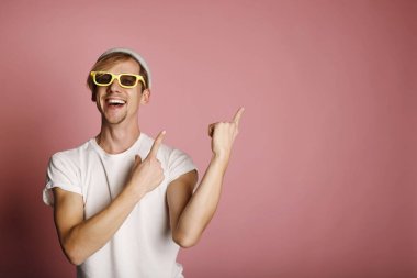 Sarı güneş gözlüklü genç adam pembe arka planda gülümsüyor ve başparmağıyla yan tarafa bakıyor. Metnin için yer