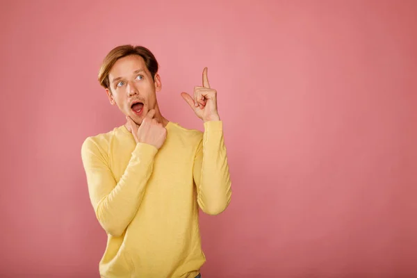 身穿黄色毛衣的年轻而快乐的男人站在粉色的背景上 脸上带着震惊的表情 用大拇指指向旁边 放在你的文字位置上 — 图库照片