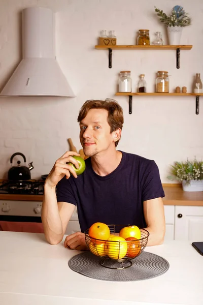 活泼的男人吃苹果时感到快乐 有魅力的年轻男性在家里的厨房里吃健康的食物 健康与减肥概念 免版税图库图片