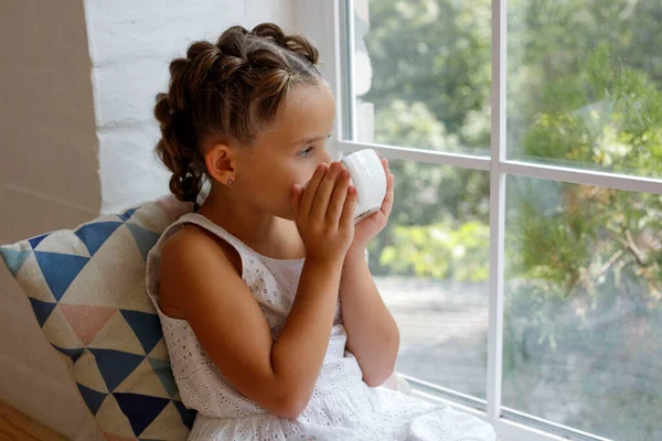 Little girl sitting by the window with a cup of hot drink and looking outdoors