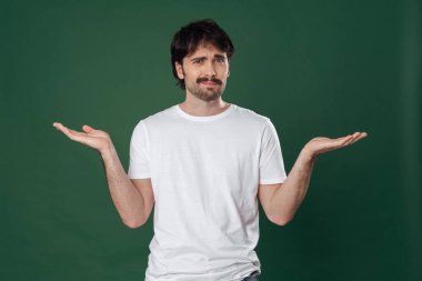 Koyu yeşil bir arka planda, beyaz tişörtlü genç bir adam kollarını yana doğru açtı ve yüzünde sorgulayıcı bir ifadeyle baktı.