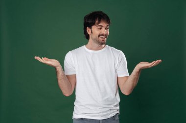 Koyu yeşil arka planda, beyaz tişörtlü genç bir adam kollarını yana açıp sağ eline bakıyor.